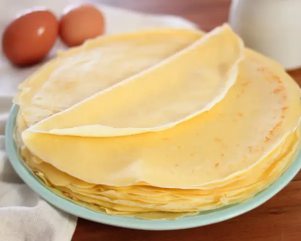 Piatto di crepes senza burro, leggere e dorate, con due uova sullo sfondo.