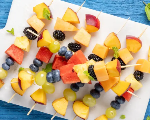 Spiedini di frutta colorati con pesche, melone, anguria, ananas, mirtilli, more e uva, guarniti con menta fresca.