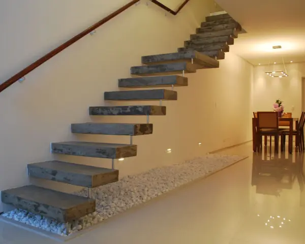 Escaleras rústicas modernas de hormigón flotante con pasamanos de madera. Un camino de guijarros blancos bordea la base.
