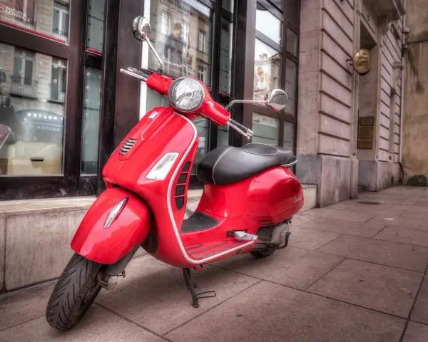 Czerwony skuter Vespa, idealny do zdobycia prawa jazdy AM, stoi na chodniku przed budynkiem.