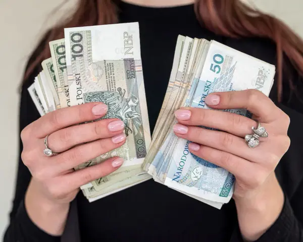 Dłonie z paznokciami w pastelowym różu trzymają pliki polskich banknotów 100 i 50 zł. To może być nagroda jubileuszowa.