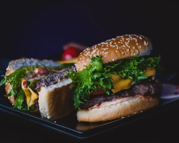 Dwa soczyste burgery z serem, sałatą i warzywami na ciemnym tle.