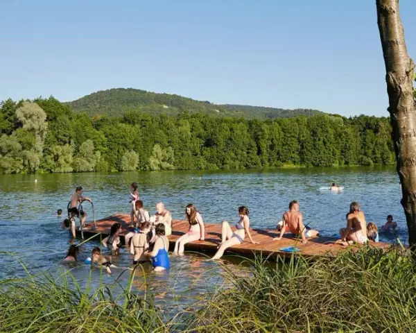 Menschen genießen einen sonnigen Tag am Baggersee Happurg. Sie sitzen auf einem Holzsteg, schwimmen und entspannen sich im Wasser.