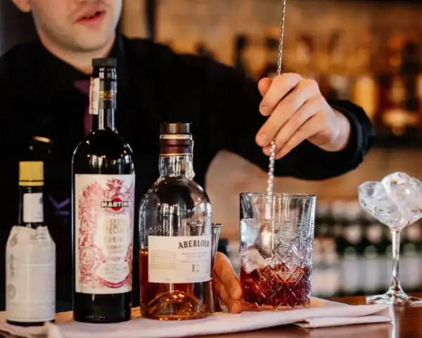 Barman miesza składniki, przygotowując najlepszy drink z whisky. Na blacie butelki Aberlour, Martini i inne.