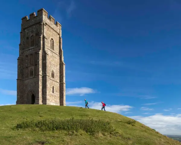 Two hikers ascend the grassy slope towards the iconic Glastonbury Tor tower under a bright blue sky.