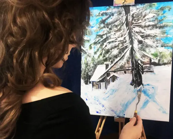 Artystka kończy malowanie zimowego domku w górach. Obraz przedstawia zasypany śniegiem krajobraz z chatką i drzewami.