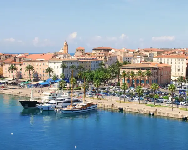 Malowniczy port na wyspie z Ajaccio, z luksusowymi jachtami i palmami, w tle kolorowe kamienice i wieża kościelna.