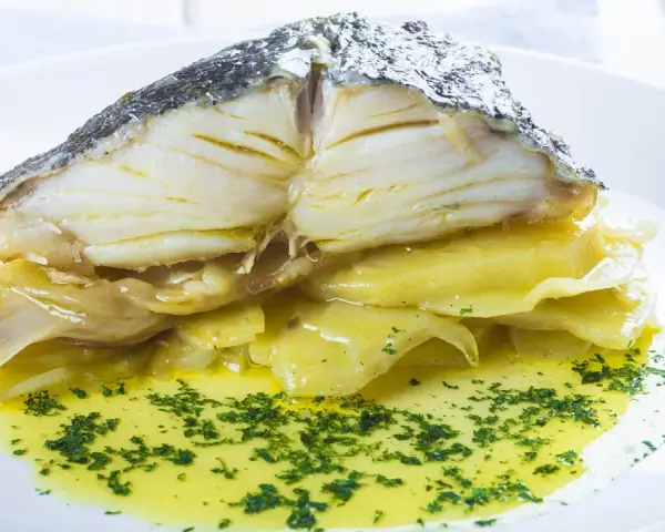 Merluza al horno con patatas, jugosa y dorada, servida sobre patatas tiernas y bañada en aceite de oliva con perejil picado.