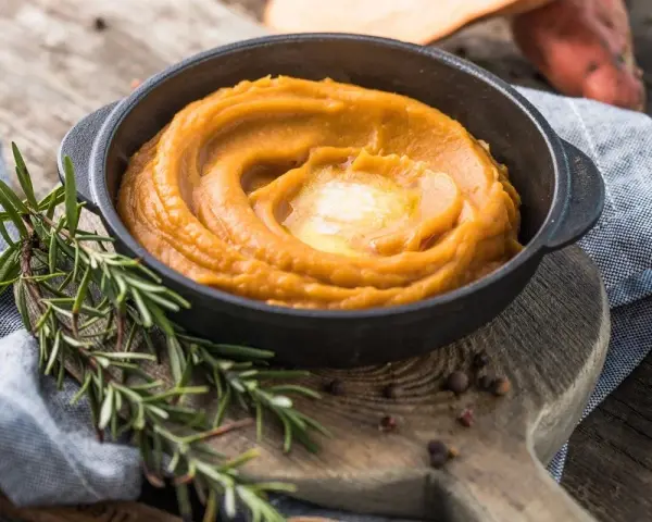 Un cremoso puré de zanahoria en un cuenco negro, coronado con mantequilla derretida. Romero fresco y granos de pimienta añaden aroma.