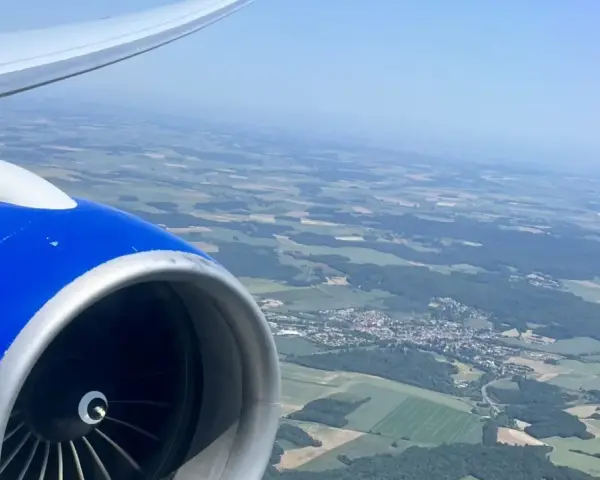 View from an airplane window, showing a blue engine and wing, with a patchwork of green fields and a small town below. Perfect for day trip flights.