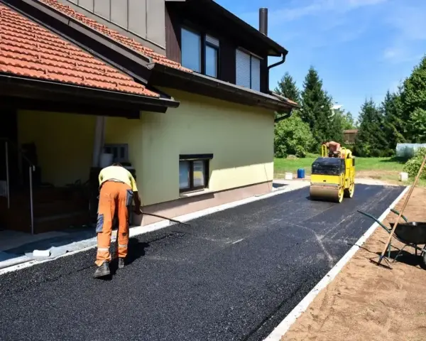Pracownik równa łopatą świeży asfalt na podjazd, a walec go ubija. Dom w tle.