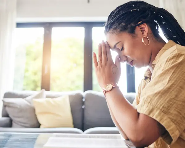A woman with braided hair and hoop earrings prays with her hands clasped, finding peace during her christian prayer times.