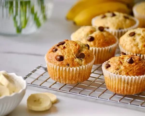 Frisch gebackene bananen muffins mit Schokostückchen auf einem Gitter, daneben eine Schüssel mit Bananenscheiben.
