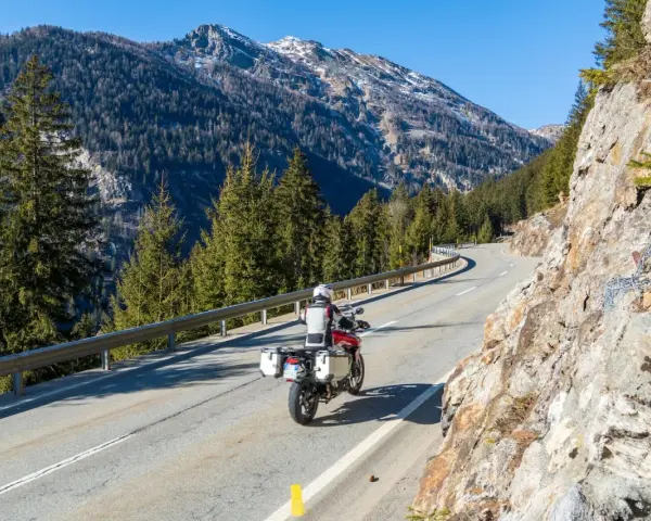 Motociclista su strada alpina in Svizzera, con montagne innevate e foreste. Ottimi consigli per un viaggio in moto in Svizzera.