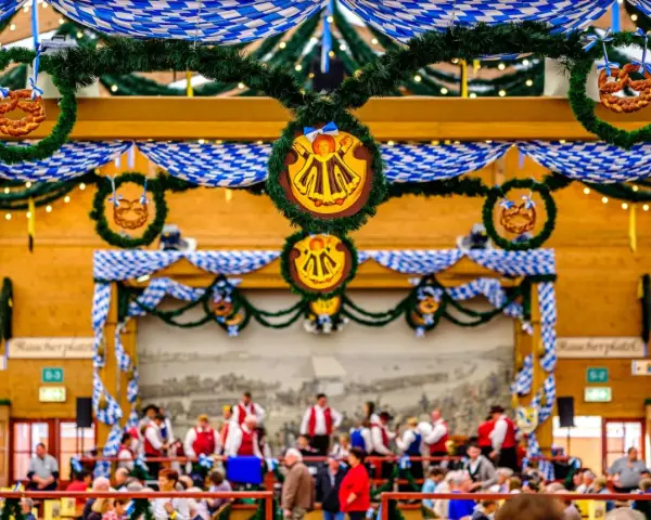Wnętrze namiotu na Oktoberfest ozdobione niebiesko-białymi girlandami i wiankami z precelkami. Ludzie siedzą przy stołach, a w tle widać orkiestrę.
