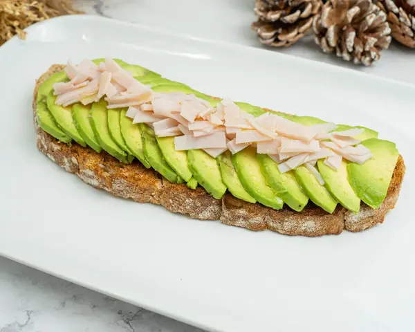 Tostada integral con aguacate en rodajas y jamón picado sobre plato blanco.