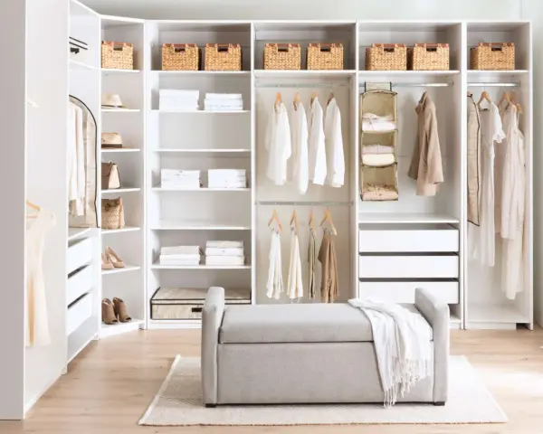 A bright, organized closet with white DIY closet shelves, featuring neatly folded clothes, baskets, and hanging garments. A grey bench sits in front.