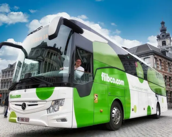 Autobus flibco.com z kierowcą, na tle miejskiej zabudowy i błękitnego nieba.