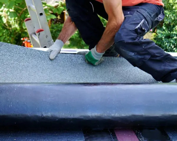 Handwerker verlegt Dachschindeln. Eine Rolle Bitumenbahn liegt bereit, während die neue Schicht auf dem Dach ausgebreitet wird.