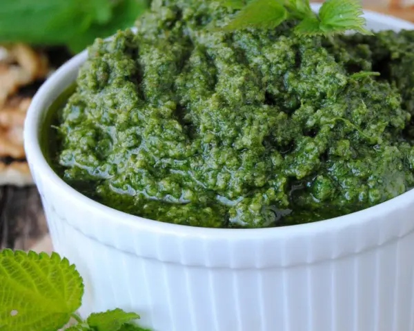 Pesto z pokrzywy w białej miseczce, obok orzechów włoskich i świeżych liści pokrzywy.