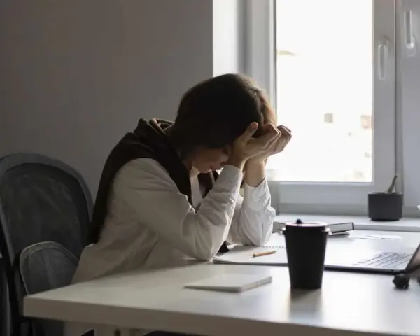 Une personne accablée par la pression au travail, la tête entre les mains, assise à un bureau encombré.