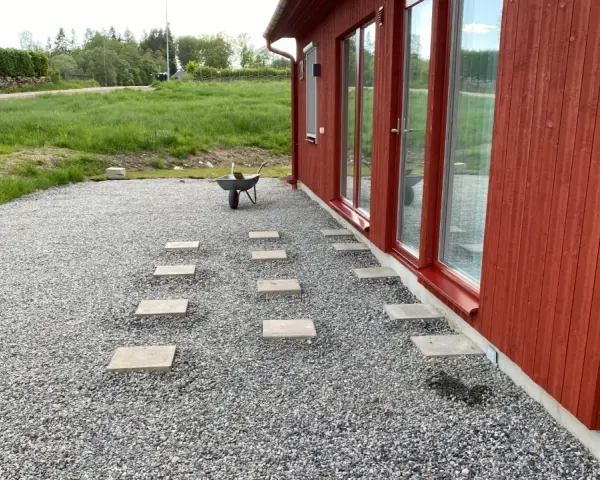 Stigande steg av altan plintar eller plattor leder genom grusgången fram till ett rött hus med stora fönster.