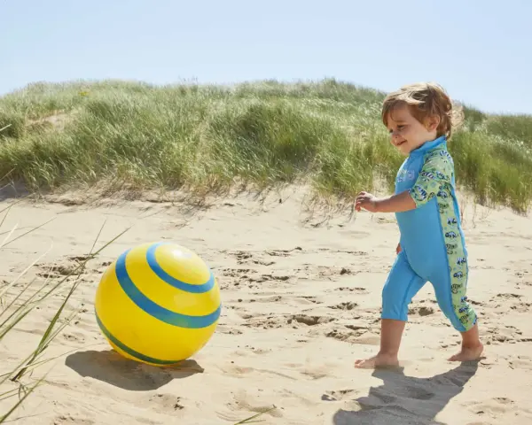 Chłopiec w niebieskim stroju kąpielowym z neoprenu bawi się żółtą piłką na plaży.