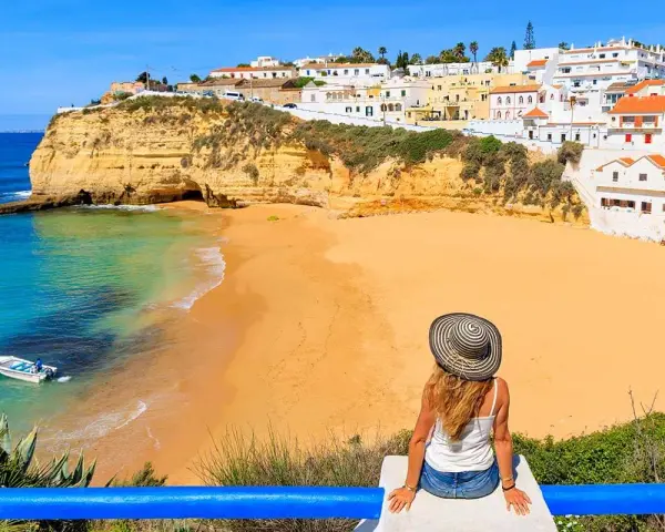 A woman in a sun hat enjoys the view of a golden beach and turquoise water in Portugal's Algarve region.
