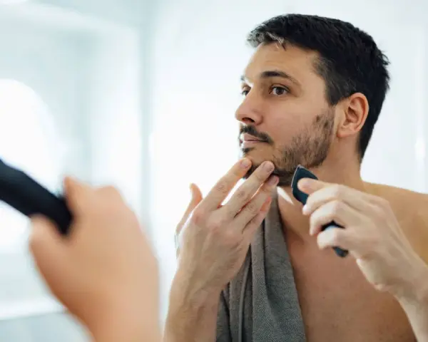 Hombre con barba corta se afeita la barba con una recortadora, buscando un afeitado apurado frente al espejo.