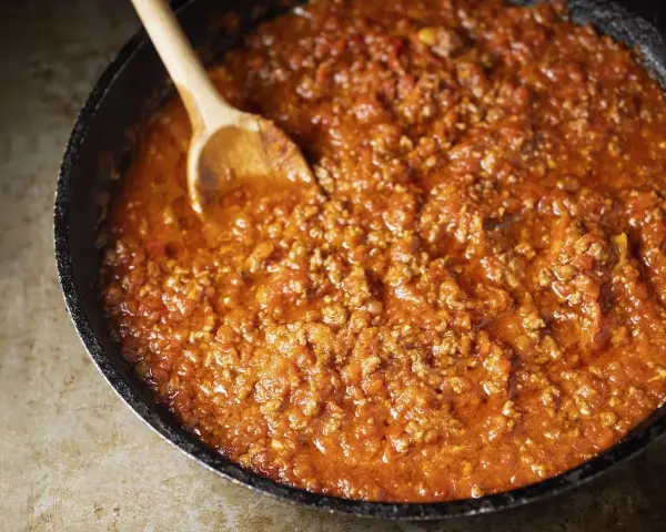Un saporito ragù di carne in una padella, pronto per essere servito. Un cucchiaio di legno mescola la salsa ricca e densa.