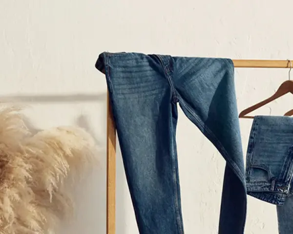 Varios pares de pantalones de **tejido denim** cuelgan de una percha de madera. A la izquierda, un gran ramo de hierba de la pampa añade un toque natural.