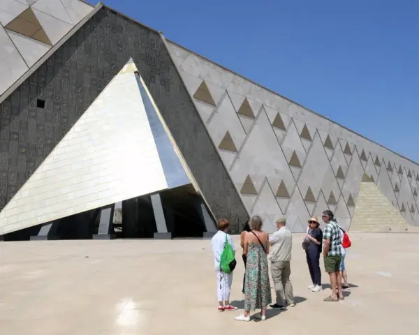Grupa turystów podziwia nowoczesne muzeum w Gizie, którego fasada zdobiona jest hieroglifami i geometrycznymi wzorami przypominającymi piramidy.