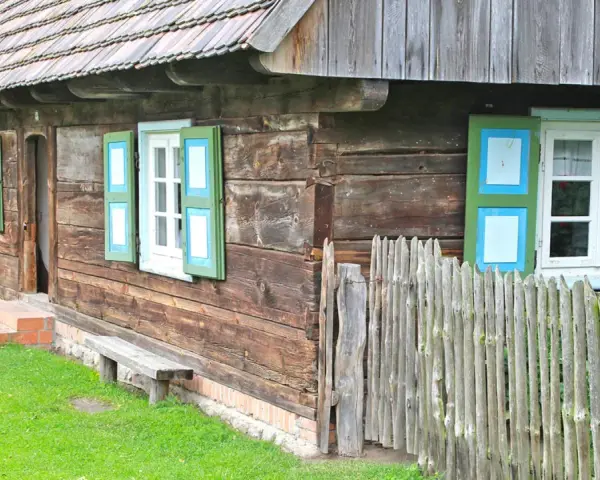 Drewniana chata z zielono-niebieskimi okiennicami, ławką i płotem. Skansen w Dziekanowicach zaprasza do podróży w czasie.
