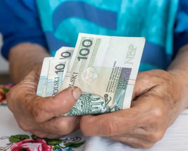 Dłonie starszej osoby trzymają banknoty 100 zł. Czy to wystarczy na godne życie, gdy średnia emerytura w Polsce jest taka, jaka jest?