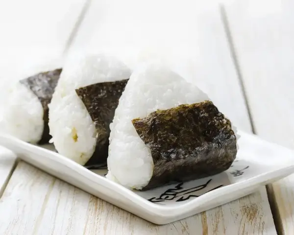 Trzy japońskie kulki ryżowe, onigiri, owinięte w ciemne wodorosty nori, ułożone na białej, ozdobnej tacce.