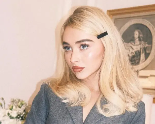 Blondynka z **side part fryzurą** i czarną spinką, w szarym żakiecie, z błękitnymi oczami.