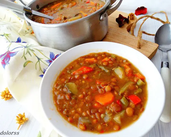 Guiso de lentejas con verduras en un cuenco blanco, junto a una olla y servilleta floral.