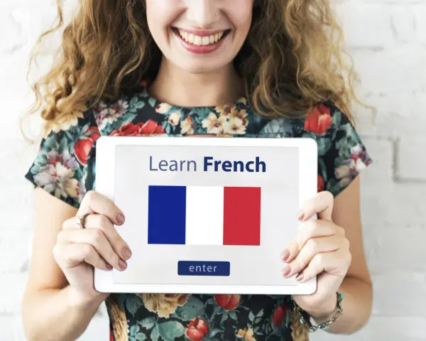 Una mujer sonriente sostiene una tablet que muestra "Learn French" y la bandera francesa. ¡Es hora de aprender como se habla en Francia!