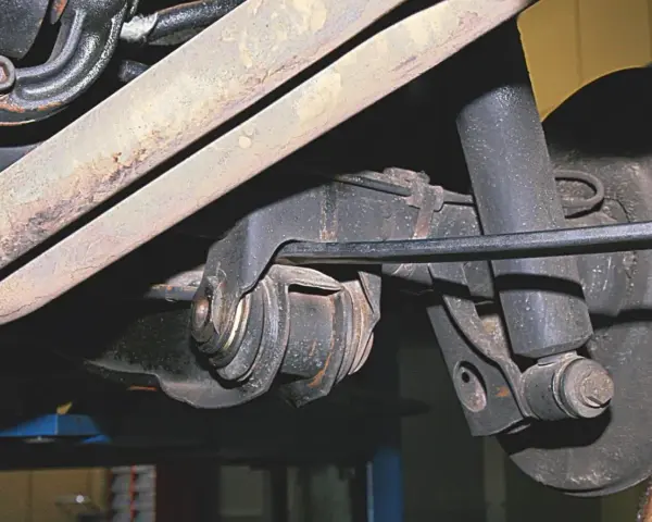 Widok z dołu na element zawieszenia samochodu. Widoczny jest metalowy wahacz z gumowymi tulejami, amortyzator i fragment układu wydechowego.