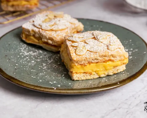 Dos porciones de pastel ruso con crema amarilla y almendras laminadas, espolvoreados con azúcar glas.