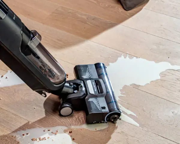 A wet/dry vacuum cleaner is cleaning up a spill of white liquid and debris on a wooden floor.