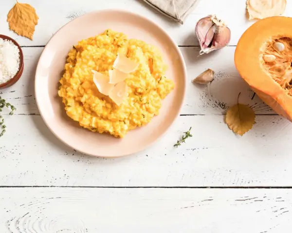 Un cremoso risotto de calabaza, decorado con láminas de queso, junto a ingredientes frescos como arroz, tomillo, ajo y una calabaza partida.