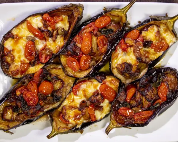 Melanzane a scarpone ripiene di pomodorini, formaggio filante e olive, cotte al forno su un piatto bianco.