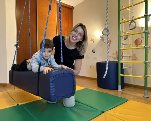 Chłopiec z ADHD u niemowlaka ćwiczy na huśtawce terapeutycznej z uśmiechniętą fizjoterapeutką.