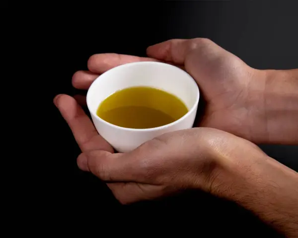 Mani che sorreggono una tazzina bianca con olio extravergine di oliva dorato. Un gesto per imparare come riconoscere olio extravergine di oliva.