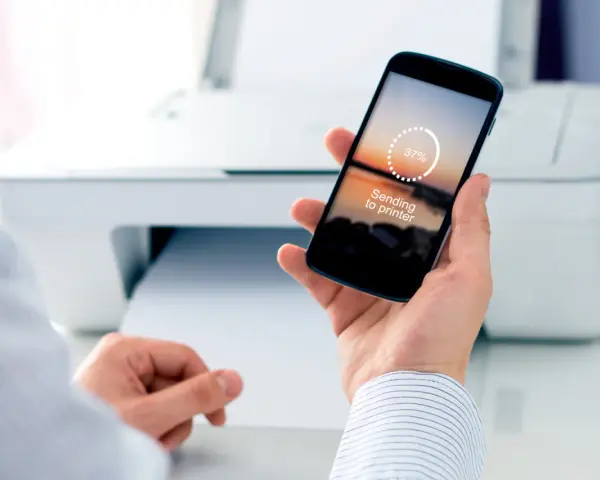 Dłoń trzyma smartfon z napisem "Sending to printer", pokazując jak drukować z telefonu. W tle biała drukarka.