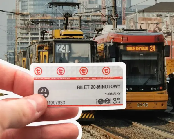 Warszawski bilet 20 minutowy w dłoni, na tle tramwajów i miejskiej zabudowy.