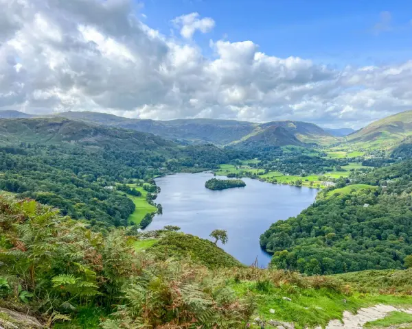 A stunning view of a lake surrounded by green hills and trees, perfect for day trips from Liverpool. A lone hiker walks down a path in the foreground.