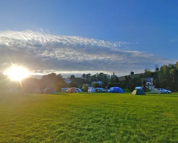 Słońce przebija się przez chmury nad polem namiotowym. Rozstawione namioty i samochody zapowiadają udany camping.