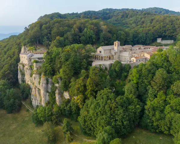 A monastery complex clings to a sheer cliff face in La Verna, Italy, surrounded by lush green forests.